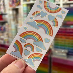 May include: A sheet of stickers featuring colorful rainbows, light blue clouds, and light blue hearts with gold trim. The rainbows have a gold outline and are in various sizes. The stickers are held in a person's hand.