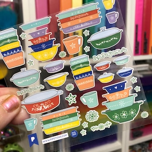 May include: A sheet of clear stickers featuring colorful vintage-style kitchenware, including bowls, mugs, and casserole dishes. The stickers have a variety of floral and geometric patterns. The sheet also includes the text "www.pipsticks.com" and "Aug 2019".