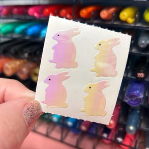 May include: A sheet of iridescent bunny stickers. The stickers are pink and yellow with a holographic finish. There are four bunnies on the sheet.