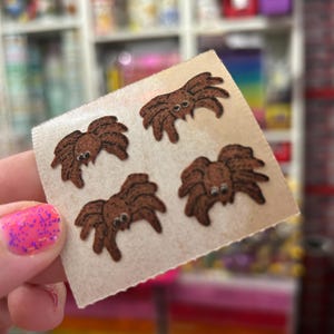 May include: A square sheet of light-colored adhesive backing featuring four small, brown, cartoon-style spider stickers. Each spider has a rounded body, eight legs, and two prominent white eyes with black pupils, arranged in a scattered pattern.