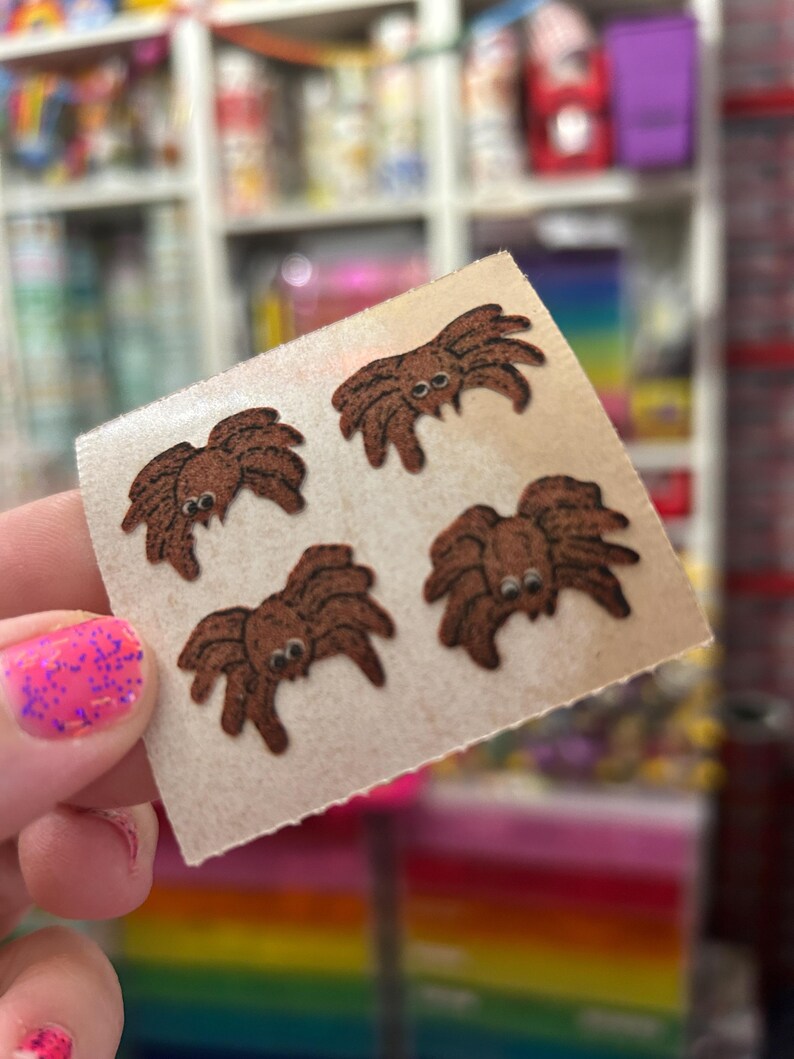 May include: A square sheet of light-colored adhesive backing featuring four small, brown, cartoon-style spider stickers. Each spider has a rounded body, eight legs, and two prominent white eyes with black pupils, arranged in a scattered pattern.