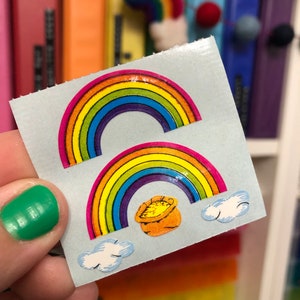 May include: Two rainbow stickers with a pot of gold at the end of the lower rainbow. The stickers are on a white background.