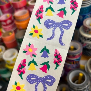 May include: A sheet of decorative stickers featuring colorful floral and bow designs. The stickers include pink, yellow, and purple flowers, along with a purple bow with white polka dots. The background is a light cream color.