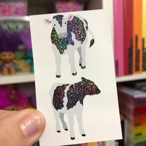 May include: Two stickers featuring cow illustrations. The cows have black and iridescent glitter patterns on their bodies, with white and gray accents. The stickers are rectangular and held by a person.