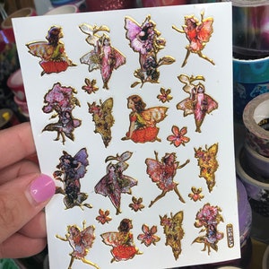 May include: A sheet of gold foil stickers featuring fairies and flowers. The stickers are on a white background and have a glossy finish.