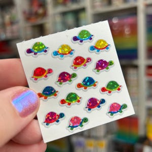 May include: A sheet of small, colorful turtle stickers. Each sticker features a turtle with a shimmering, iridescent shell in various colors, including green, yellow, red, and blue. The turtles have black eyes and are set against a white background.