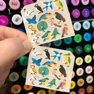May include: Two sheets of stickers featuring various bird illustrations. The stickers are on a beige background and feature a variety of birds, including a peacock, a puffin, a swan, and a robin. The stickers are arranged in a grid pattern.