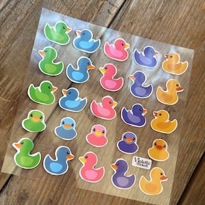 May include: A sheet of colorful rubber duck stickers. The ducks are in various colors, including green, blue, pink, yellow, and purple. The stickers are arranged in rows on a clear sheet. The sheet has the text "Violette Stickers" in the bottom right corner.
