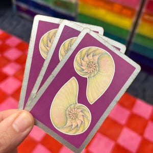 May include: Three tarot cards with a purple background and a silver border. Each card features a yellow nautilus shell design. The cards are held in a hand, with a red and pink checkered surface in the background.