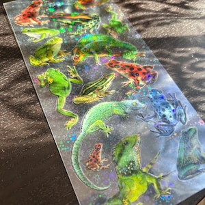 May include: A sheet of iridescent stickers featuring various frogs, lizards, and other amphibians. The stickers are on a clear background with a holographic sparkle effect. The stickers are perfect for adding a touch of whimsy to any project.