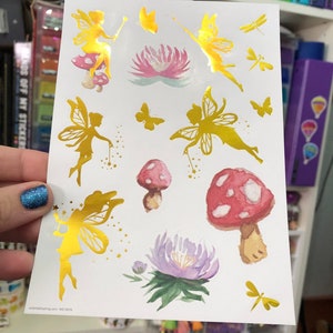 May include: A sheet of gold foil stickers featuring fairies, mushrooms, flowers, and dragonflies. The stickers are on a white background.