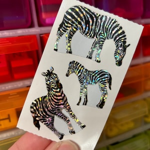 May include: A sheet of three holographic zebra stickers. The zebras have black and silver stripes and are in different poses. The stickers are on a white background.