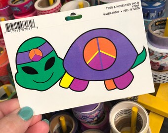 May include: A sticker sheet with a green alien wearing a purple headband and a purple turtle with a peace sign on its shell. The sticker sheet is labeled "Water-Proof Peel 'N' Stick" and "#7061".