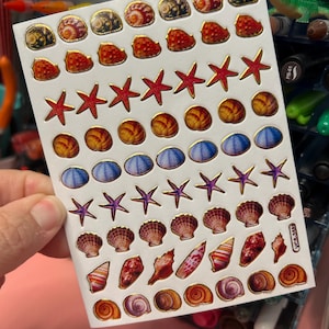 May include: A sheet of colorful stickers featuring various sea-themed designs, including seashells, starfish, and ocean-inspired patterns. The stickers are arranged in rows on a white background.
