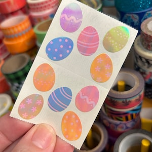 May include: A sheet of colorful iridescent Easter egg stickers. The stickers feature various designs, including polka dots, wavy lines, and flowers.