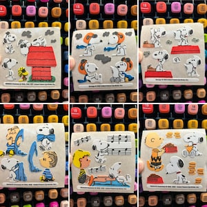 May include: A set of nine vintage Peanuts character sticker sheets. Each sheet features a different scene with characters like Snoopy, Charlie Brown, and Woodstock. The stickers are printed on white paper with a light brown background.