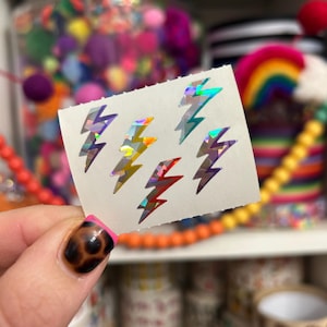 May include: A sheet of six holographic lightning bolt stickers in various colors, including purple, yellow, and red. The stickers are held up against a blurred background of colorful craft supplies.