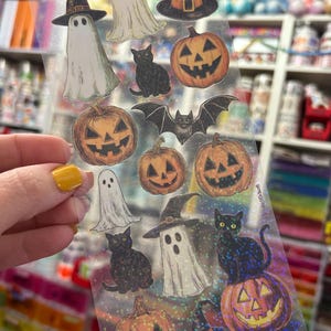 May include: A sheet of Halloween-themed stickers featuring ghosts wearing witch hats, jack-o'-lanterns, black cats, and a bat. The stickers have a holographic, glittery finish, and are in various sizes and shapes. The overall color scheme is orange, black, and white.