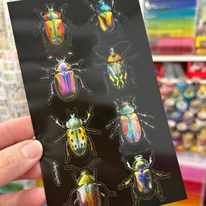 May include: A sheet of black stickers featuring detailed illustrations of various iridescent beetles. The beetles have a variety of colors, including gold, blue, green, and red. The stickers are held by a person's hand.