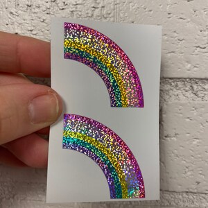 May include: Two rainbow-colored, glittery stickers in a semi-circular shape. The stickers are on a white background and held by a hand. The rainbow colors include red, orange, yellow, green, and blue.
