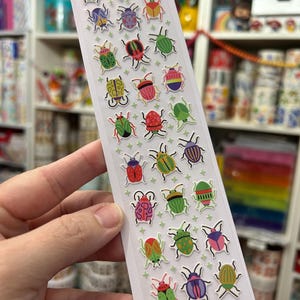 May include: A vertical sticker sheet featuring a collection of colorful, cartoon-style beetle illustrations in various patterns and hues, arranged in a grid with small green cross symbols. The bottom includes the text BRIGHT BEETLES WWW.PIPSTICKS.COM.