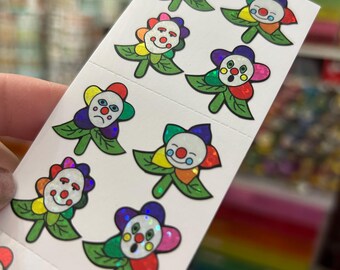 May include: A sheet of stickers featuring colorful flower designs with clown faces. Each flower has a different expression, with petals in rainbow hues and green leaves. The stickers have a holographic, glittery effect.