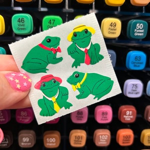 May include: A sheet of stickers featuring four green frogs with different accessories. The frogs are wearing hats, bow ties, and a tie. The stickers are on a white background.