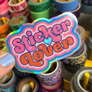 May include: A holographic sticker with the words "Sticker Lover" in pink and orange bubble letters. The sticker has a cloud-like border with a blue, pink, and orange colour scheme. The background features various rolls of decorative tape.
