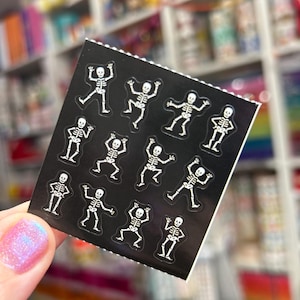 May include: A sheet of black stickers featuring white skeleton illustrations in various poses. The stickers are held in a hand with a pink and purple glitter nail polish. The background is a blurred image of shelves.