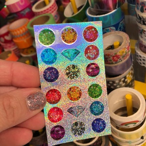 May include: A sheet of holographic stickers featuring various colorful gemstone designs. The stickers include round and diamond-shaped gems in shades of green, orange, red, blue, and purple. The background is a shimmering, glittery silver.