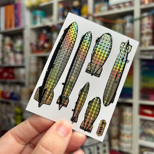 May include: A sheet of holographic stickers featuring various airship designs. The stickers have a metallic sheen with black and gold accents. The designs include several zeppelin shapes and a small rectangular sticker with the number "1849-B".