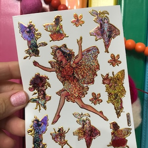 May include: A sheet of gold glitter fairy stickers with a variety of poses and designs. The fairies are wearing red, gold, and purple dresses with iridescent wings. The stickers are on a white background.