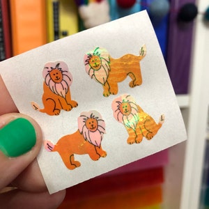 May include: Four iridescent orange lion stickers with black outlines on a white sheet of paper. The lions have pink manes and are facing forward.