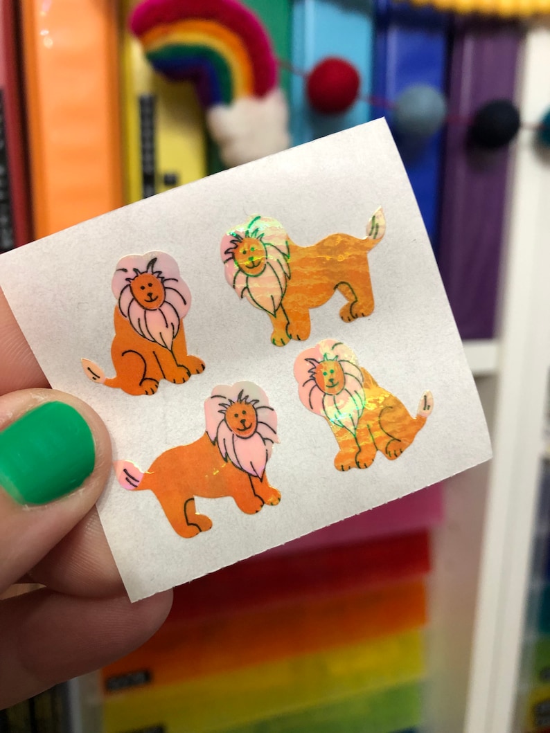 May include: Four iridescent orange lion stickers with black outlines on a white sheet of paper. The lions have pink manes and are facing forward.
