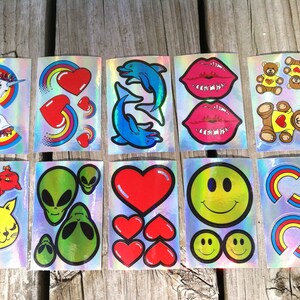 May include: A sheet of holographic stickers featuring various designs, including rainbows, hearts, dolphins, lips, teddy bears, aliens, and smiley faces.