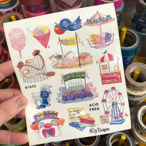 May include: A sheet of stickers featuring a variety of carnival and fairground themes. The stickers include a Ferris wheel, a clown, a pie, a pig race, a ring toss, a cotton candy stand, a corn dog stand, and a crazy cups game. The stickers are printed on white paper with a blue border. The text on the stickers includes "Fun House", "Crazy Cups", "Pig Races", "Ring Toss", "Corn Dogs", "Ferris Wheel", "Acid Free", and "NON Designs".