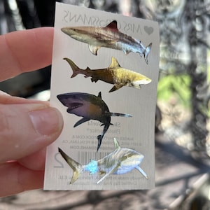 May include: A sheet of four silver holographic shark stickers. The stickers are on a white background with the text "Mrs. Grossman's" and "Photo Stickers" printed on the sheet.