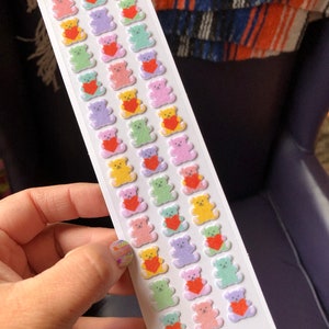 May include: A sheet of colorful fuzzy teddy bear stickers with red hearts. The stickers are arranged in rows on a white background. The sheet has the text "Fuzzy Stuffed Bears" and "www.pinspicks.com" printed on it.