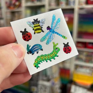 May include: A sheet of colorful insect stickers. The stickers include a ladybug, a bee, a dragonfly, a caterpillar, and a beetle. The stickers have a shiny, glittery finish and are in various colors, including red, yellow, green, and blue.