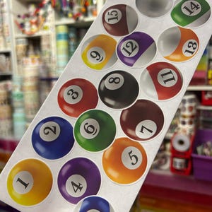 May include: A rectangular sticker sheet featuring fifteen circular billiard ball designs, each labeled with a unique number from one to fifteen, including solid and striped patterns in various colors like yellow, blue, red, purple, orange, green, brown, and black.