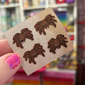 May include: A square piece of light brown cardstock featuring four small, brown, fuzzy spider-shaped stickers. Each spider has a rounded body, multiple legs, and two small, white, circular eyes with black pupils, arranged in a grid pattern.