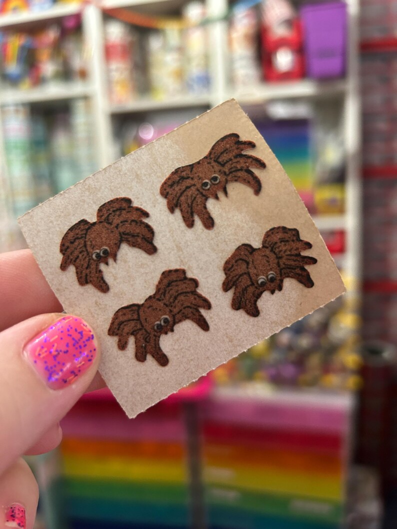 May include: A square piece of light brown cardstock featuring four small, brown, fuzzy spider-shaped stickers. Each spider has a rounded body, multiple legs, and two small, white, circular eyes with black pupils, arranged in a grid pattern.