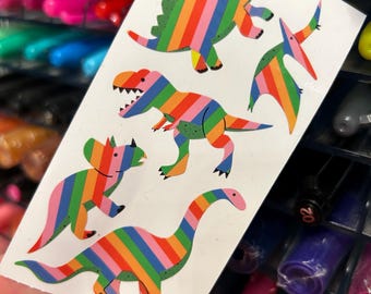 May include: A sheet of colorful dinosaur stickers with a rainbow stripe pattern. The stickers include a stegosaurus, a tyrannosaurus rex, a triceratops, and a brachiosaurus.