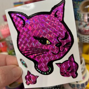May include: A sheet of holographic stickers featuring a pink cat with a winking eye and a pearl necklace. Three smaller pink cat stickers are also included.
