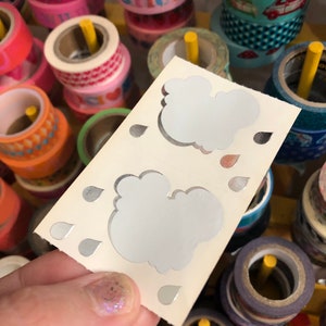 May include: A white card with two silver cloud stickers and silver raindrop stickers. The card is held in a hand against a background of colorful washi tape rolls.
