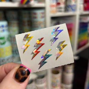 May include: A sheet of six holographic lightning bolt stickers in yellow, red, silver, and teal. The stickers have a reflective, iridescent finish. The stickers are on a white background.