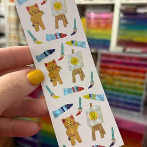 May include: A sheet of stickers featuring a repeating pattern of a cartoon cat artist, a smiling sun on an easel, paint tubes, and paintbrushes. The stickers have a pearlescent sheen and are held by a hand with yellow nail polish.