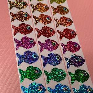 May include: Strips of colorful, glittery fish stickers in various colors, including gold, orange, red, purple, blue, green, and teal. Each fish has a small, round eye. The stickers are on white backing.