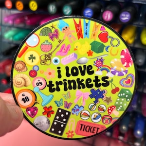 May include: A green circle sticker with the text "i love trinkets" in a retro font. The sticker is filled with various colorful trinkets, including a troll doll, a key, a horseshoe, a butterfly, a domino, a ticket, and a puzzle piece.