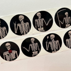 May include: Eight round stickers featuring black and white skeleton illustrations on a black background. The skeletons are in various poses, including holding a scythe, a light bulb, and a rose.